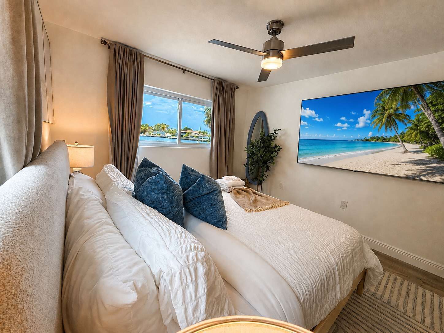 Habitación de hotel con cama, ventilador de techo, ventana con cortinas, planta y pintura con vista al mar.