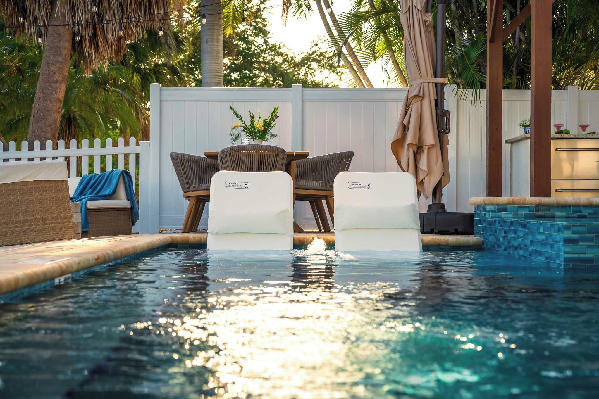 A swimming pool with two chairs and a table, surrounded by a white fence and palm trees.