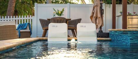 A swimming pool with two chairs and a table, surrounded by a white fence and palm trees.