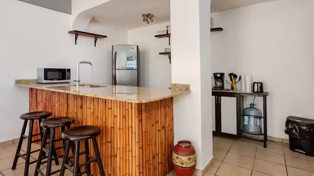 Uma cozinha com ilha central, banquetas, micro-ondas, geladeira e bebedouro.