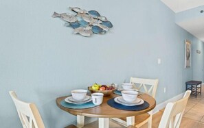 A dining area with a round table set for four, surrounded by white chairs. A fruit bowl is placed on the table, and there is a wall-mounted decorative piece above it. - Direct Garage Access, Lake View & 5 Pools | Laketown Wharf 226 (Panama City Beach)