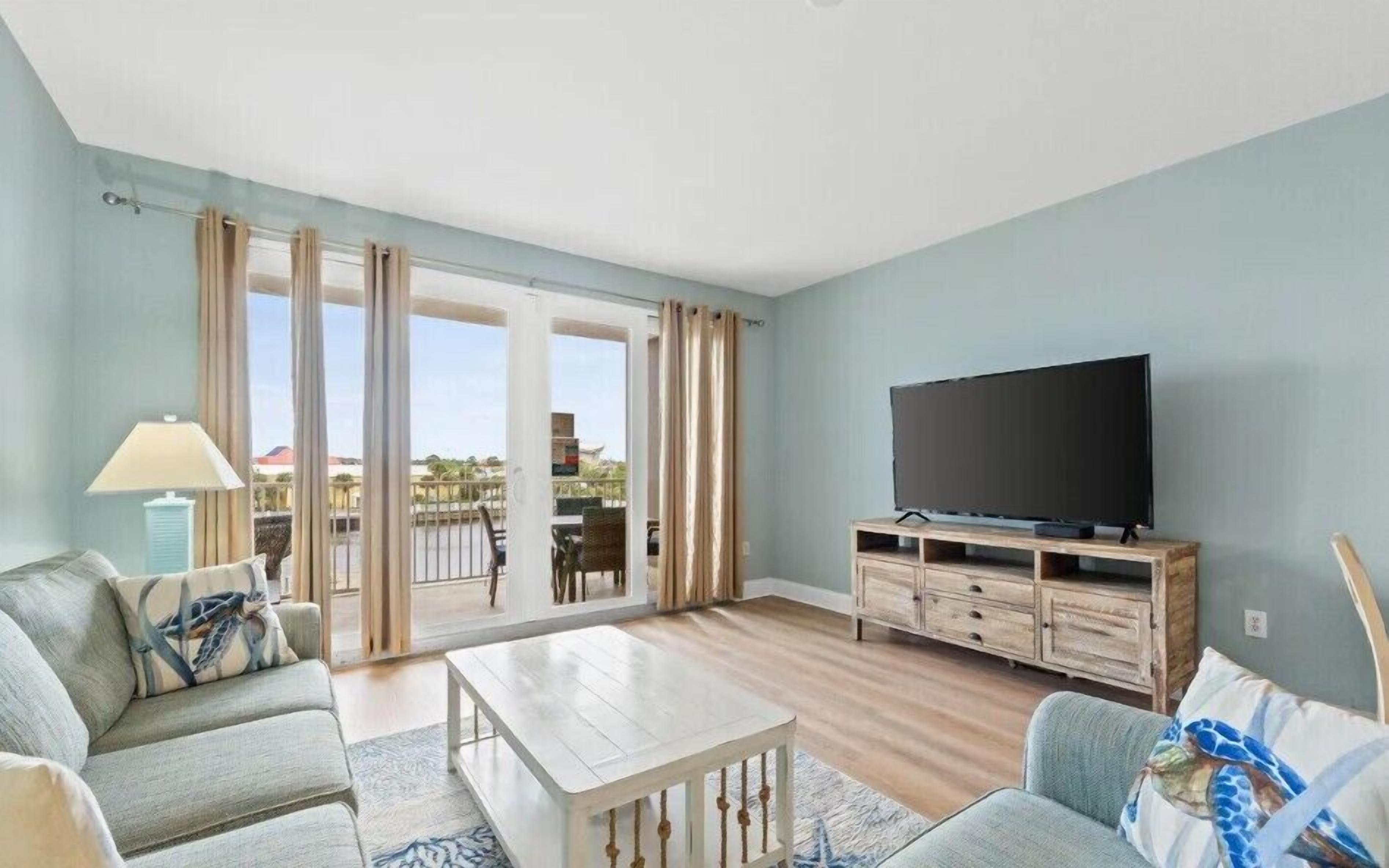 Ein Wohnzimmer mit Sofa, Couchtisch, Fernseher auf einem Holzgestell und Blick durch eine Glastür auf einen Außenbereich.