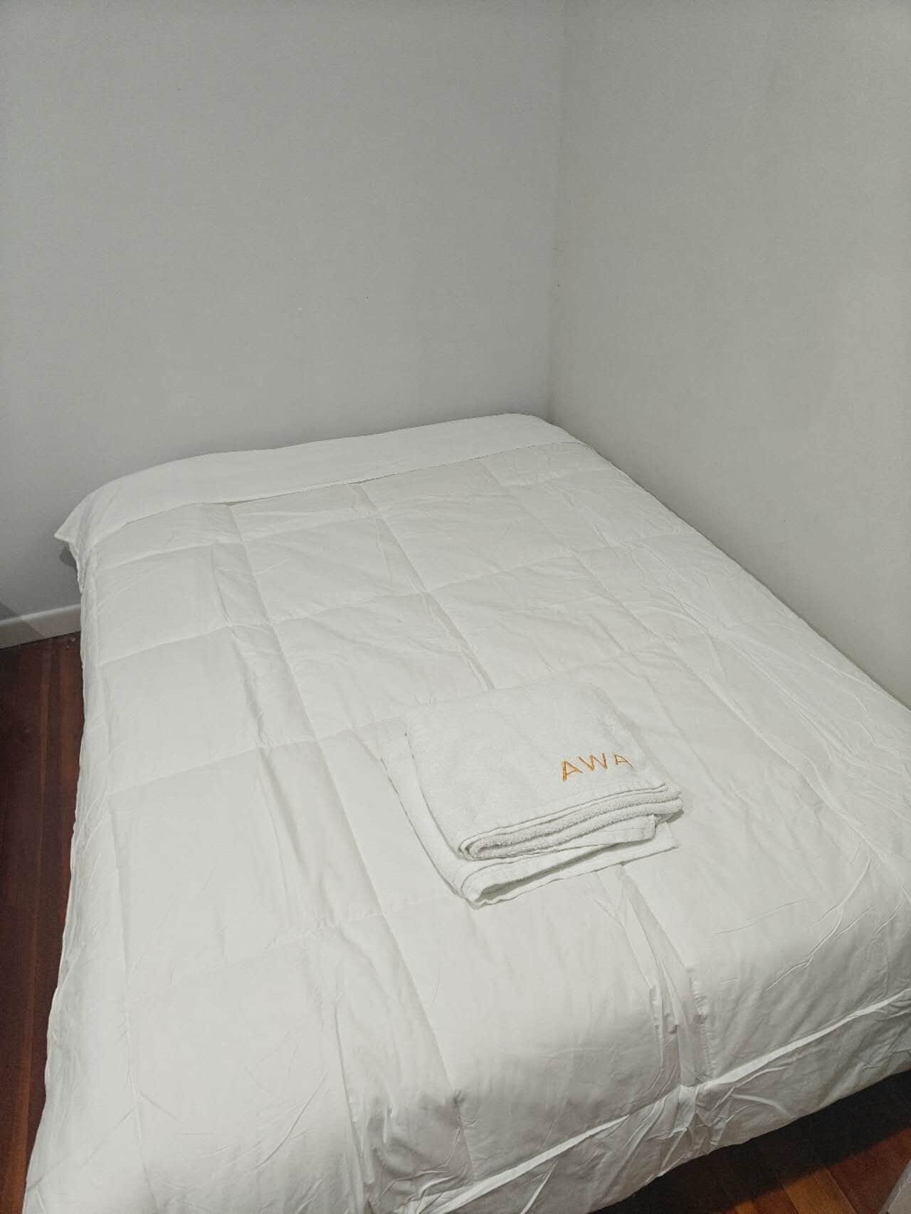 Ein Einzelbett mit weißer Daunendecke und einem gefalteten Handtuch mit 'AWA' aufgestickt.