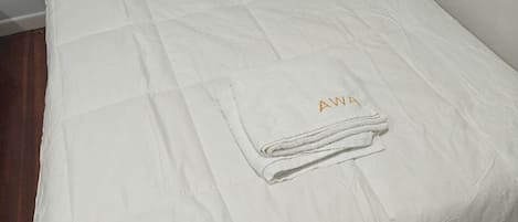 Una cama individual con edredón blanco y una toalla doblada con el bordado 'AWA'.