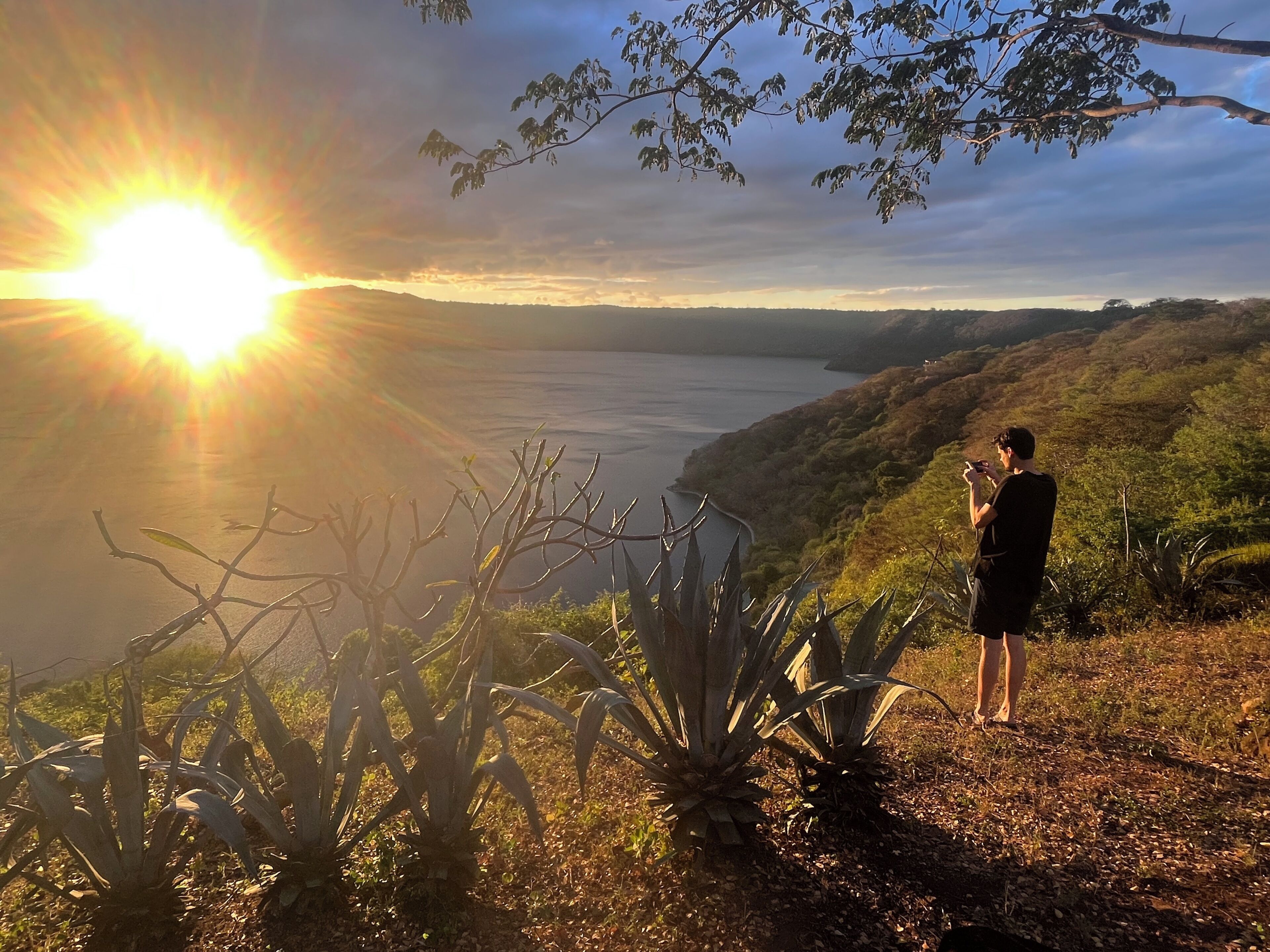 Una persona está de pie en una colina con vistas a un cuerpo de agua y un atardecer de fondo.