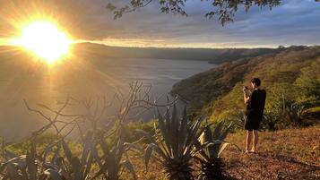 Una persona está de pie en una colina con vistas a un cuerpo de agua y un atardecer de fondo.