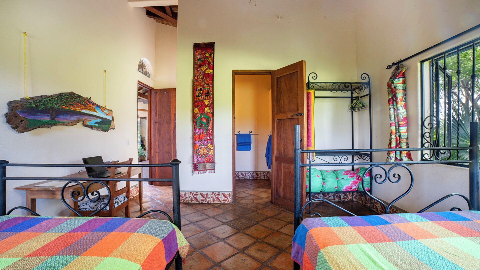 Une chambre avec deux lits, un bureau en bois, une décoration murale colorée et une vue sur l’extérieur par une fenêtre.