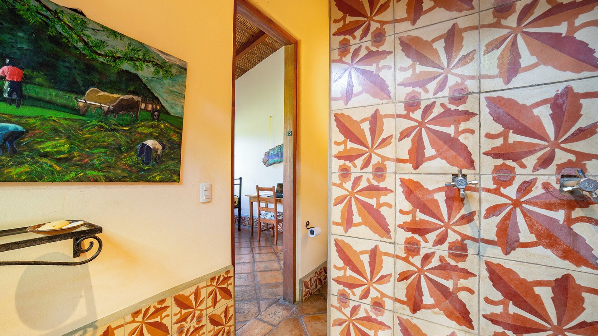 A hallway with a tiled wall featuring a leaf pattern, a painting of a rural scene, and a metal rack with a plate.