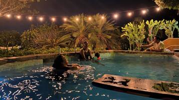 A swimming pool area with people, surrounded by greenery and string lights.