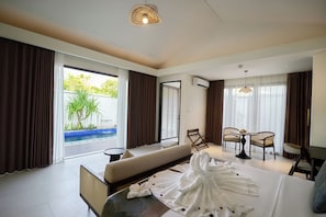 A hotel room with a bed, a sofa, a dining table with chairs, a small balcony with a pool, and a view of greenery outside.