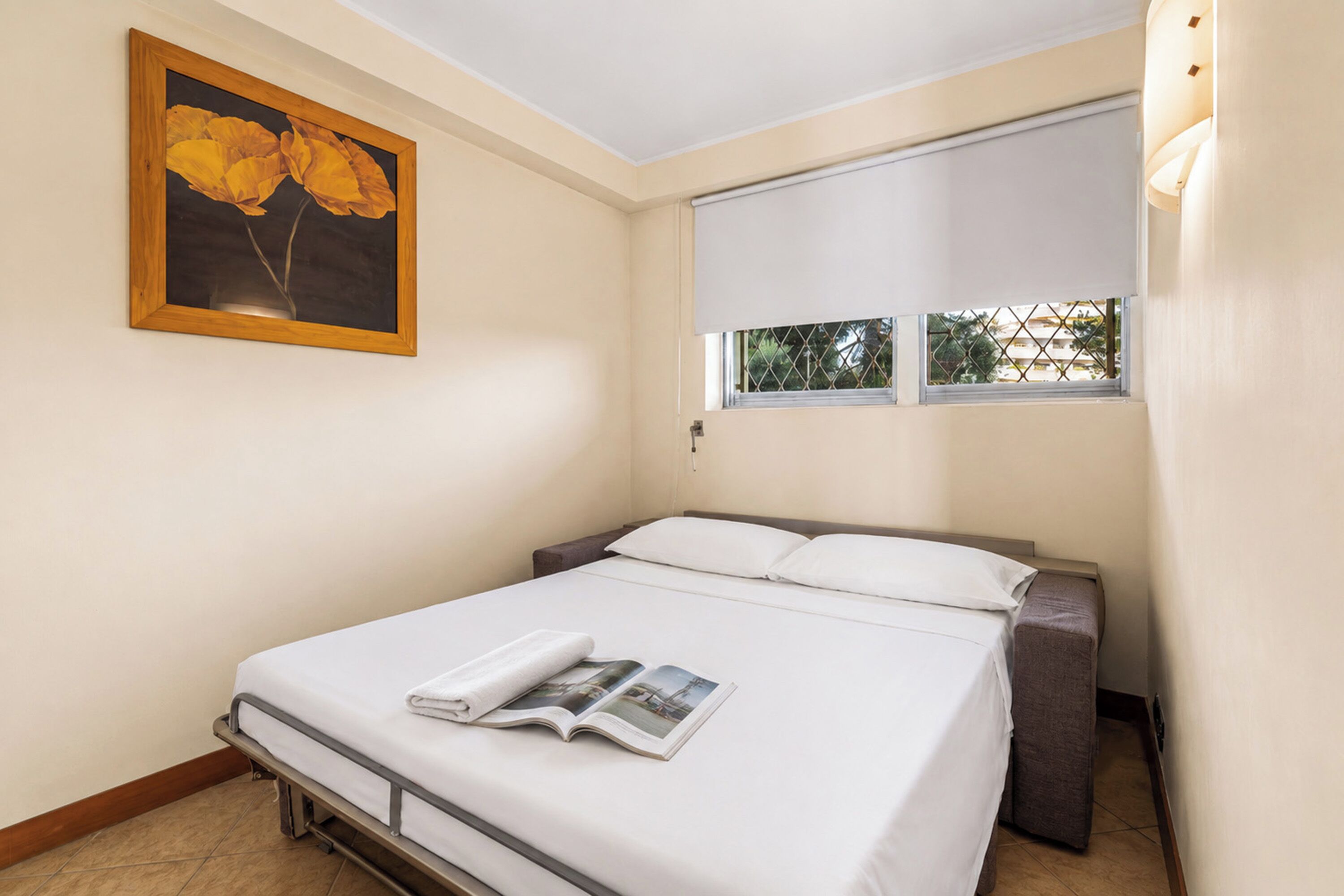 A hotel room with a bed, a window with a view, a painting on the wall, and a magazine on the bed.