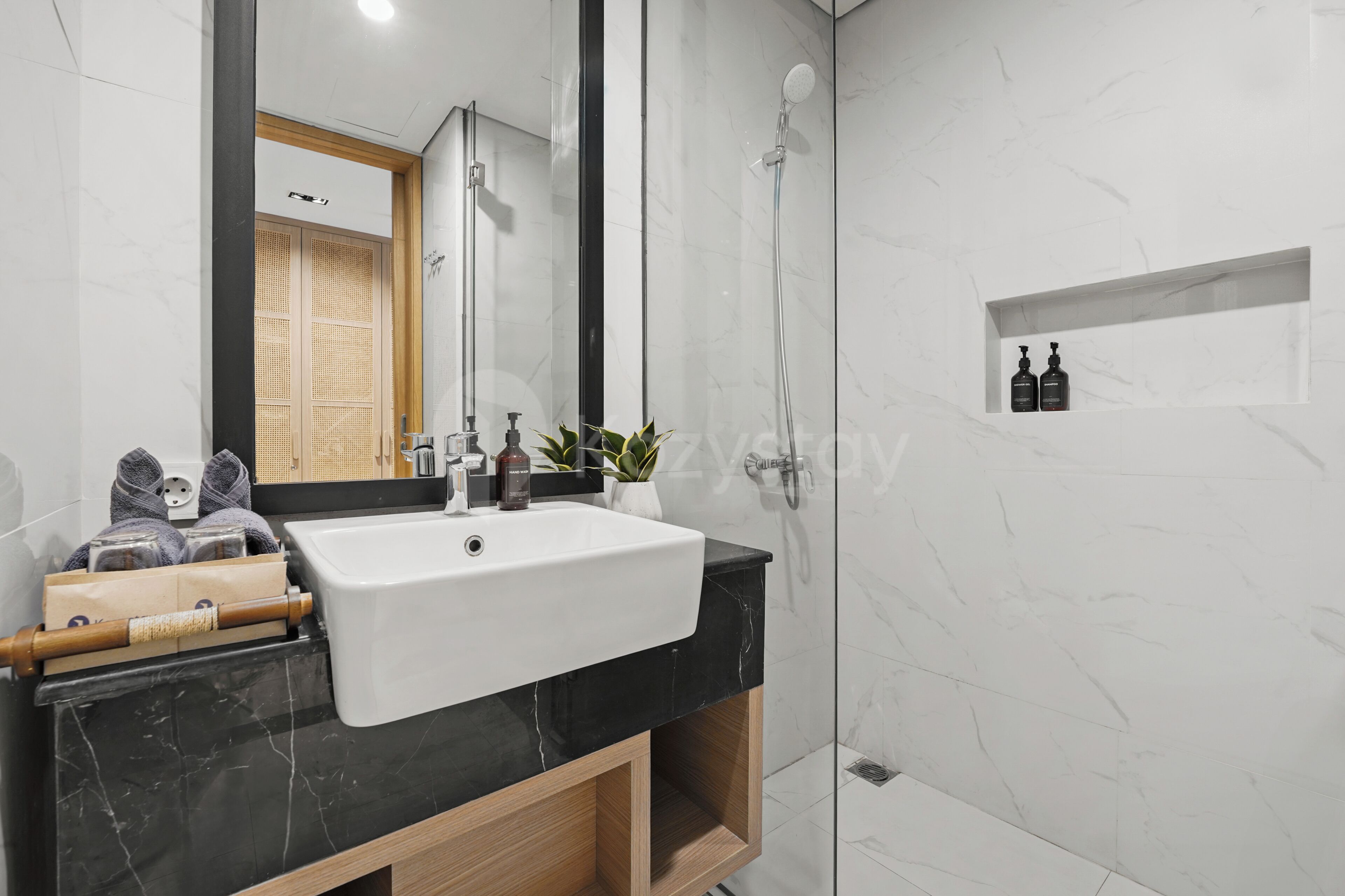 A modern bathroom with a white sink, a mirror, a shower, and a shelf with toiletries.