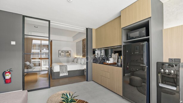 Una habitación de hotel moderna con una cama, una mesa de comedor con una planta, una zona de cocina con refrigerador y microondas, y un extintor visible.