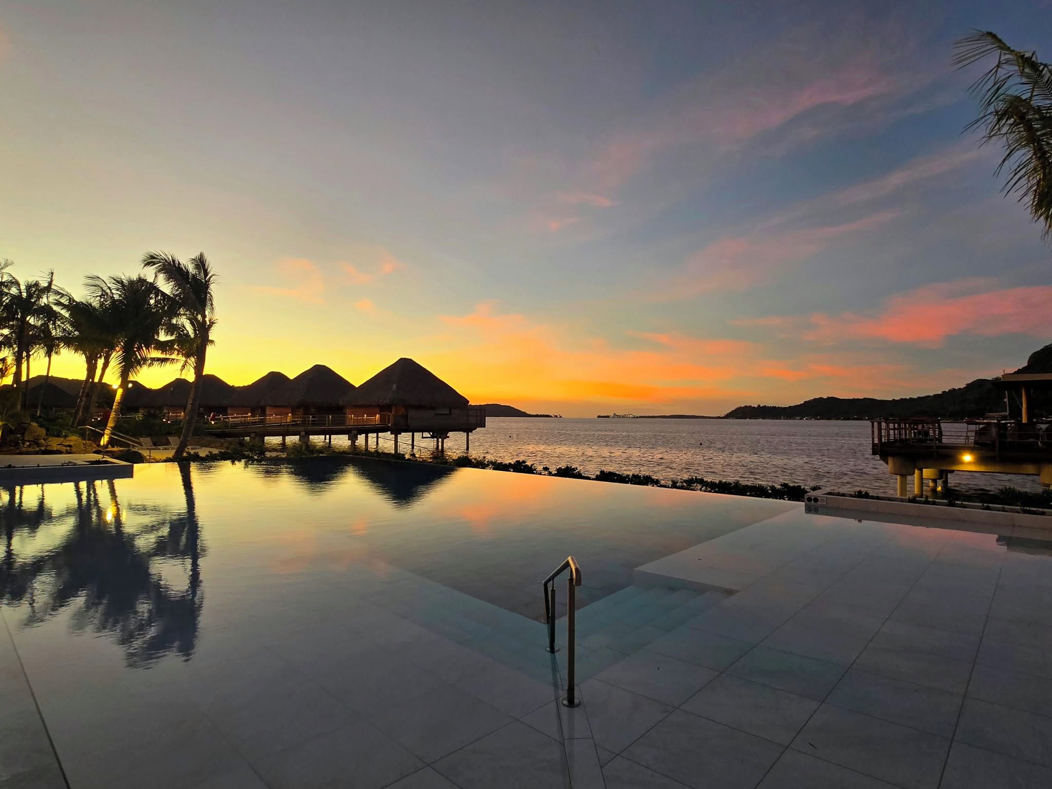 An infinity pool with a clear view of the sunset over water, surrounded by overwater bungalows and palm trees.