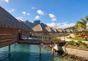 Overwater bungalows with thatched roofs, a wooden walkway over water, and a clear blue sky.