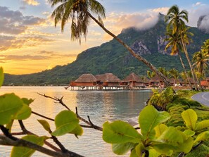 Overwater bungalows with thatched roofs, a calm sea, and a mountainous backdrop.