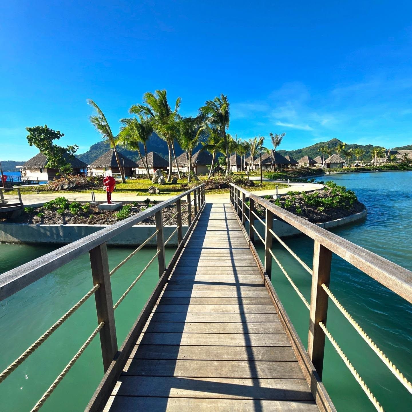 A wooden bridge leading to a resort area with thatched-roof buildings and palm trees.