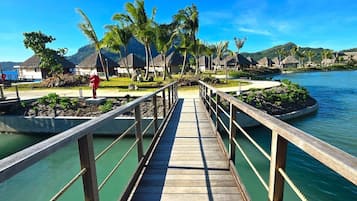 A wooden bridge leading to a resort area with thatched-roof buildings and palm trees.