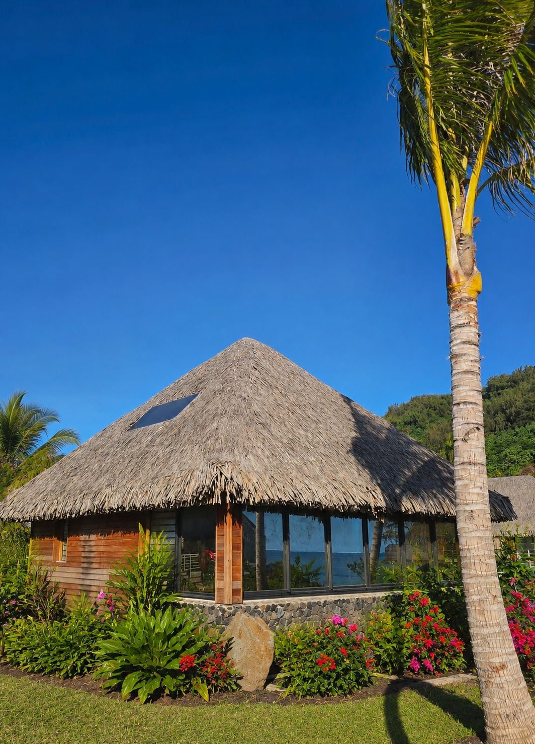 Un bâtiment à toit de chaume, avec une façade en bois, entouré d’une végétation tropicale et surplombé d’un ciel bleu azur.