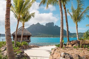 A beach with a thatched-roof hut, palm trees, and a mountain in the background. - Bloody Mary's Hotel (Bora Bora)