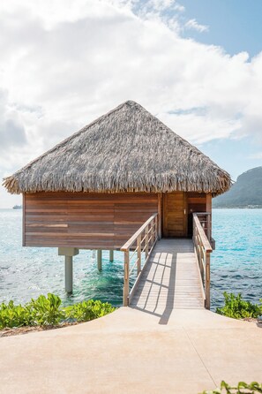 An overwater bungalow with a thatched roof, wooden walls, and a wooden walkway leading to the entrance. - Bloody Mary's Hotel (Bora Bora)