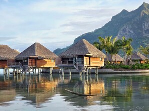 Overwater bungalows with thatched roofs, wooden structures, and a calm water body reflecting the buildings.