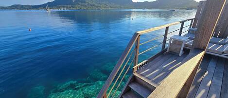 A wooden deck with a railing overlooking clear blue water and a mountainous landscape.