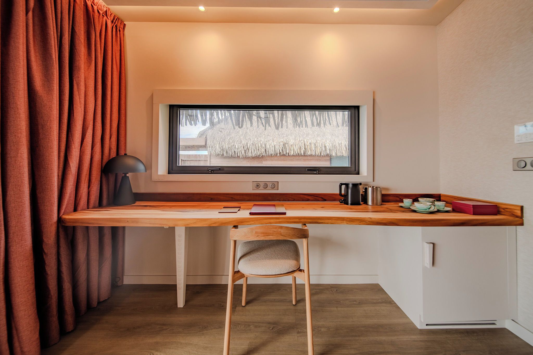 Habitación pequeña con un escritorio de madera, una silla, una ventana que da a una cabaña de paja, una lámpara negra y un hervidor.