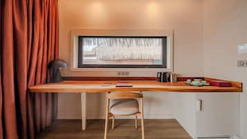 Habitación pequeña con un escritorio de madera, una silla, una ventana que da a una cabaña de paja, una lámpara negra y un hervidor.