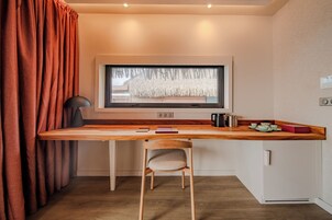 Habitación pequeña con un escritorio de madera, una silla, una ventana que da a una cabaña de paja, una lámpara negra y un hervidor.