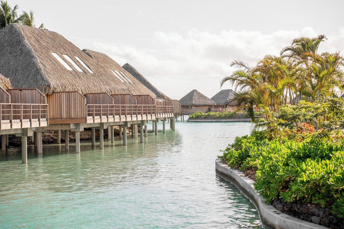 Overwater bungalows with thatched roofs, connected by walkways over calm turquoise water.