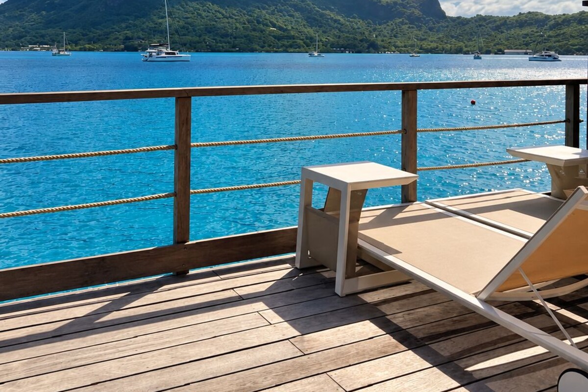 A wooden deck with lounge chairs and tables overlooking a body of water with boats and a mountain in the distance.