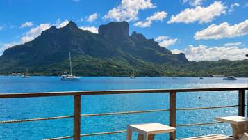 A wooden deck with lounge chairs and tables overlooking a body of water with boats and a mountain in the distance.