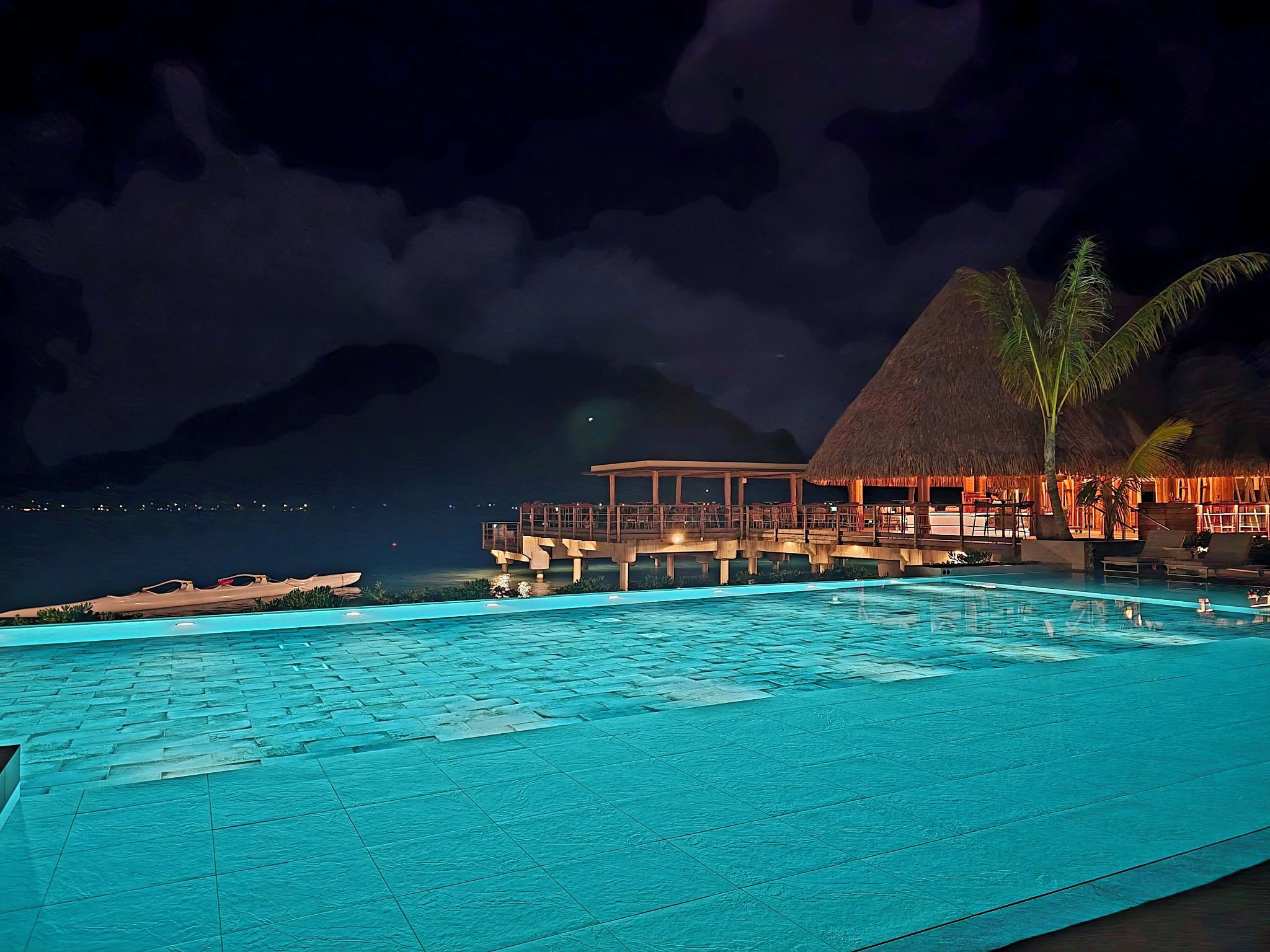 A nighttime view of a resort with a swimming pool, a thatched-roof structure, and a mountain in the background.