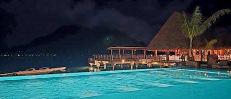 A nighttime view of a resort with a swimming pool, a thatched-roof structure, and a mountain in the background.