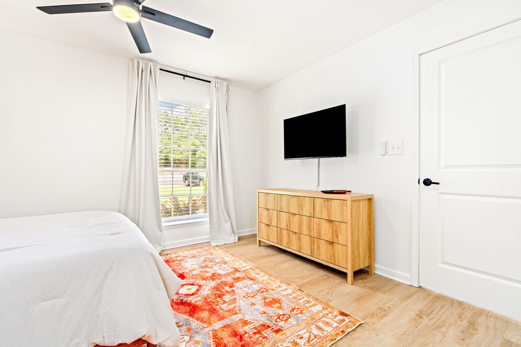 A bedroom with a bed, a TV mounted on the wall, a wooden cabinet, and a window with curtains.