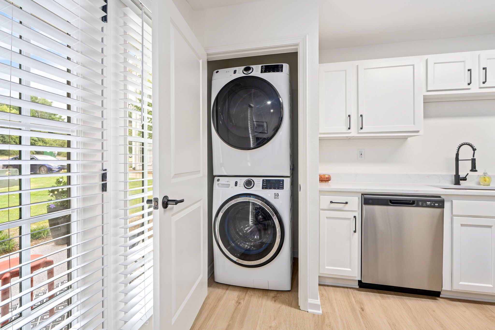 A laundry room with a front-loading washer and dryer, white cabinets, and a view through a door with blinds.