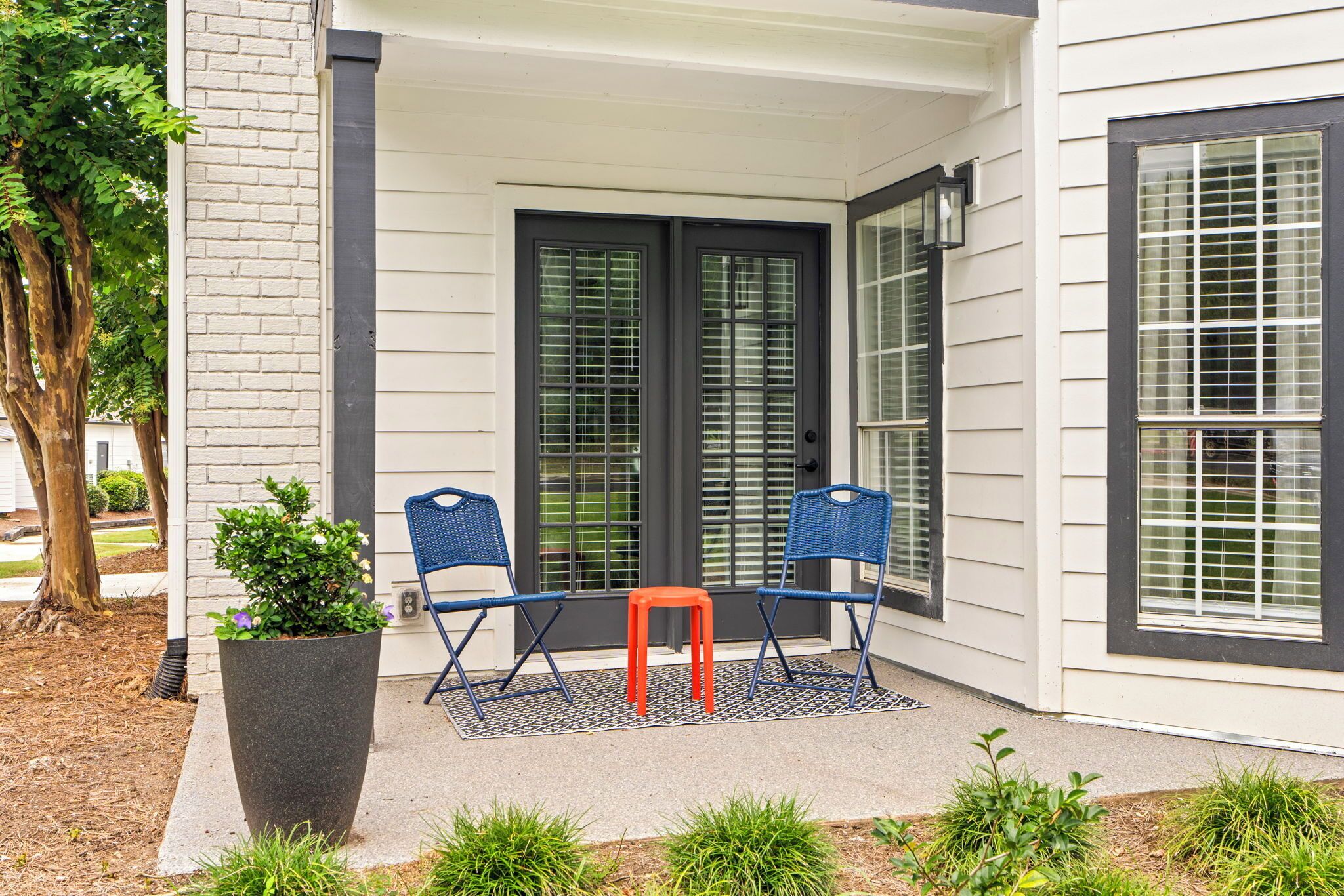 Une terrasse couverte avec deux chaises bleues et un petit tabouret orange, une plante en pot et un tapis à motifs.