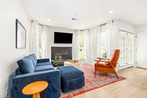 Una sala moderna con un sofá azul, una mesa de centro de madera, una alfombra roja estampada, una chimenea y un televisor montado en la pared.