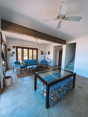 A spacious living area with a glass coffee table, blue sofas, and a ceiling fan. - Wake up to the sea | 3 suites | Monte Alto Beach (Arraial do Cabo)