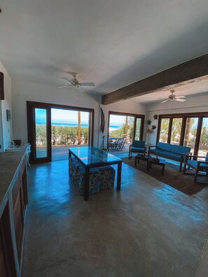 A spacious living area with a glass-top table, a sofa, and a bench. There are ceiling fans and large windows offering a view of the outdoors. - Wake up to the sea | 3 suites | Monte Alto Beach (Arraial do Cabo)