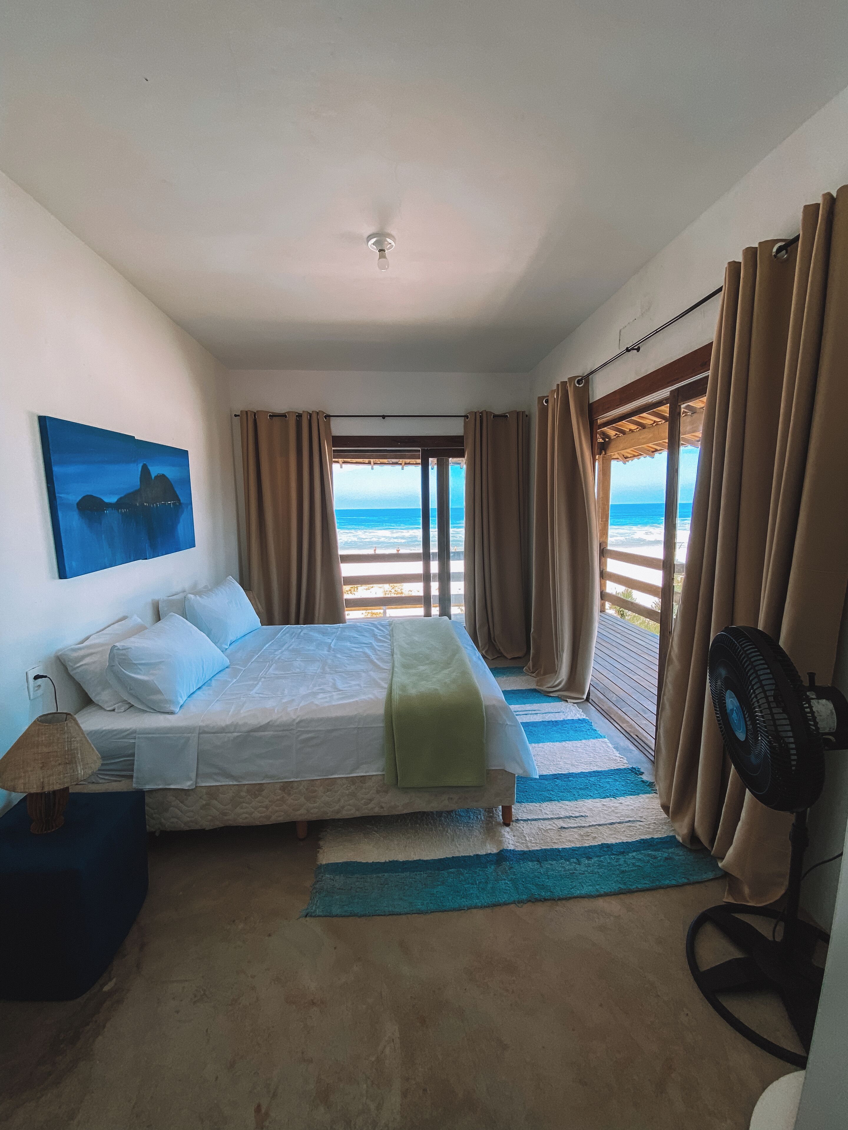 A bedroom with a bed, a fan, a lamp, a painting, and a view of the ocean.