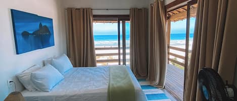 A bedroom with a bed, a fan, a lamp, a painting, and a view of the ocean.