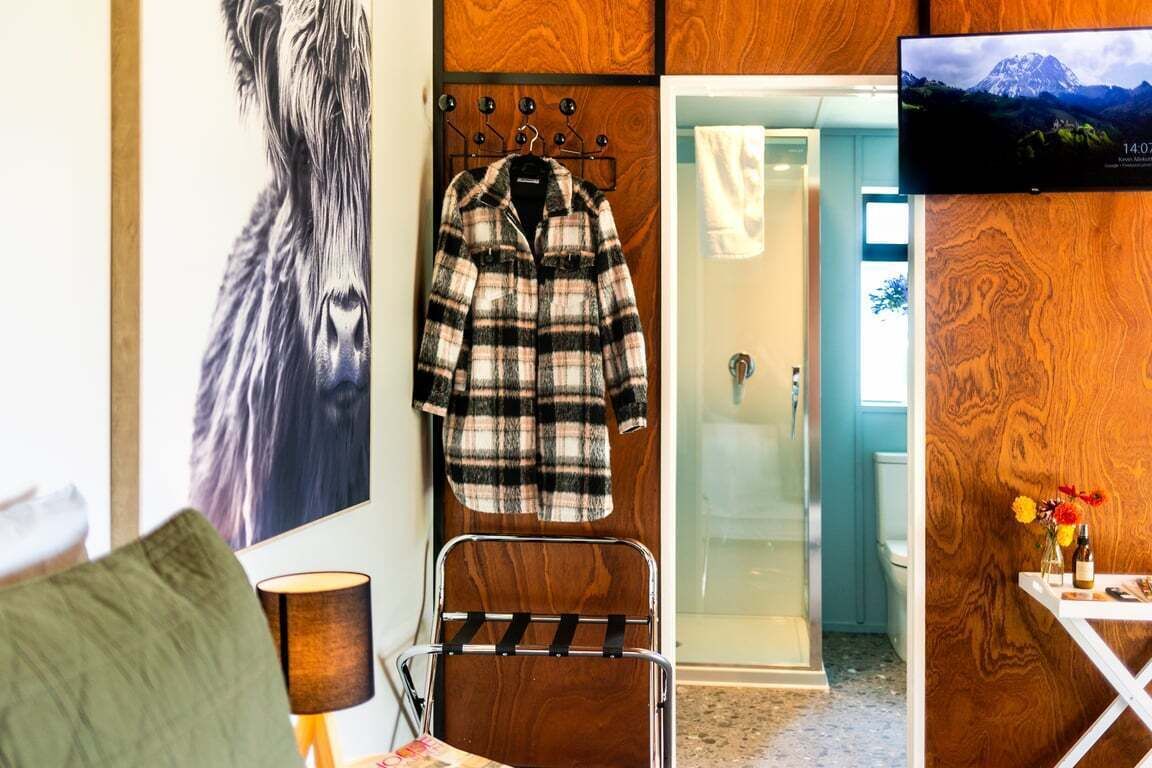 A hotel room with a wooden wall, a coat rack with a plaid jacket, a TV screen displaying a mountain landscape, and a small table with a vase of flowers.