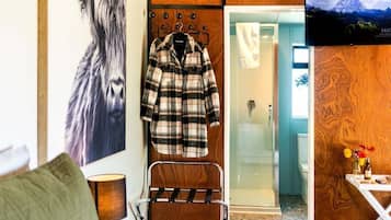 A hotel room with a wooden wall, a coat rack with a plaid jacket, a TV screen displaying a mountain landscape, and a small table with a vase of flowers.