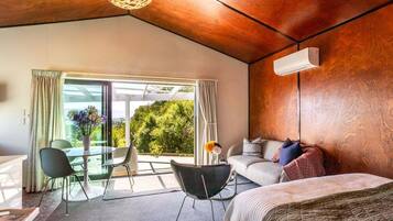 A modern hotel room with a wooden ceiling, a sofa, a bed, a dining area with chairs, and a large window offering a view of greenery.