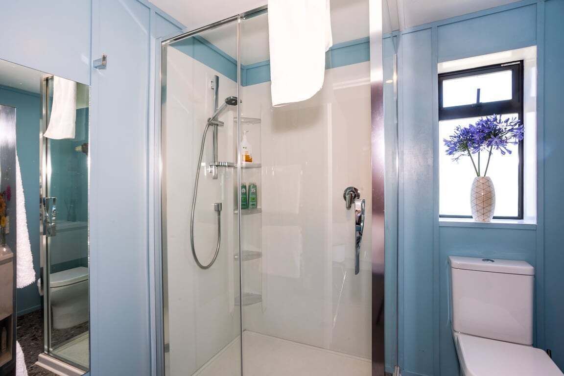 A bathroom with a glass-enclosed shower, a toilet, and a window with a vase of flowers.