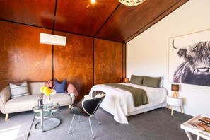 Une chambre moderne avec un plafond en bois, un lit recouvert d’une couverture verte, un canapé avec des coussins, une table basse en verre et un climatiseur fixé au mur.