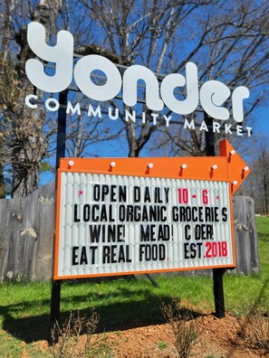 Placa do Yonder Community Market, indicando que está aberto diariamente das 10h às 18h, com mantimentos orgânicos locais, vinho, carne e comida de verdade, fundado em 2018.