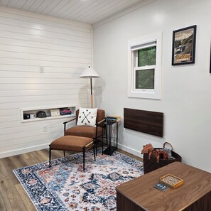 Um quarto com piso de madeira, uma cadeira marrom, um tapete estampado, uma pequena mesa com um jogo de tabuleiro e uma prateleira fixada na parede com livros e objetos decorativos.